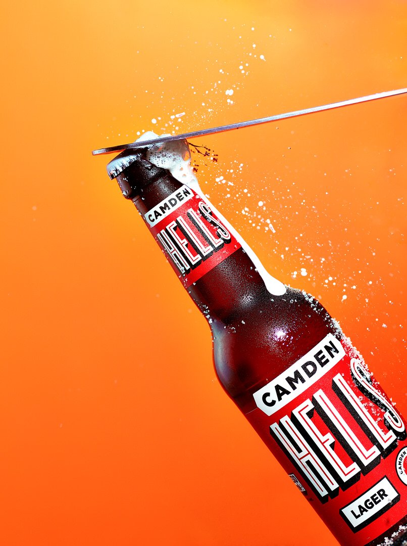 Bottle of Camden Hells lager being opened, with foam splashing over an orange background