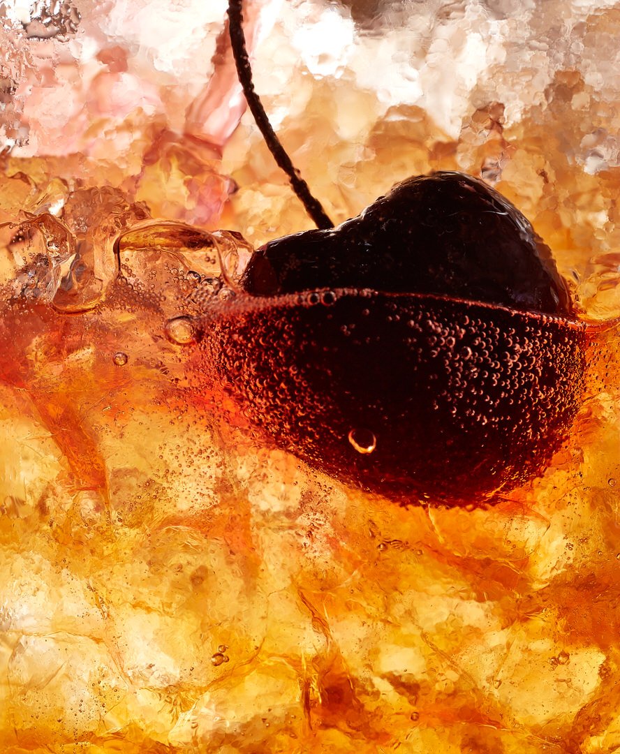 Close-up of a cherry with its stem submerged in a fizzy, amber-coloured drink, covered in tiny bubbles