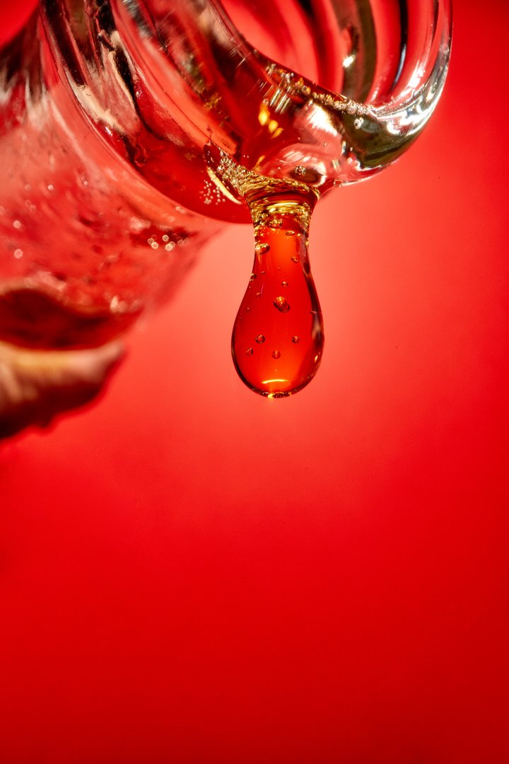 Close-up of a golden liquid droplet forming at the neck of a bottle against a red background