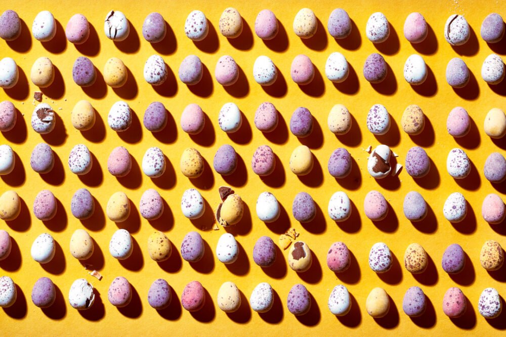 Rows of speckled mini chocolate eggs in pastel colours arranged in a grid on a yellow background, some cracked open revealing chocolate