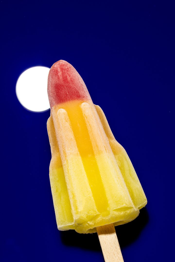 Rocket-shaped ice lolly with a red tip and yellow body, set against a dark blue background with a white moon-like circle