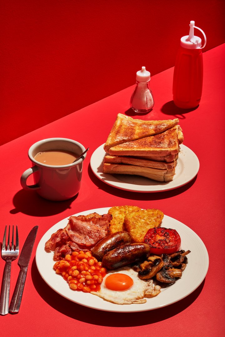 Full English breakfast with bacon, sausages, egg, beans, hash browns, mushrooms and tomato, served with toast, tea and sauce bottles on a red background