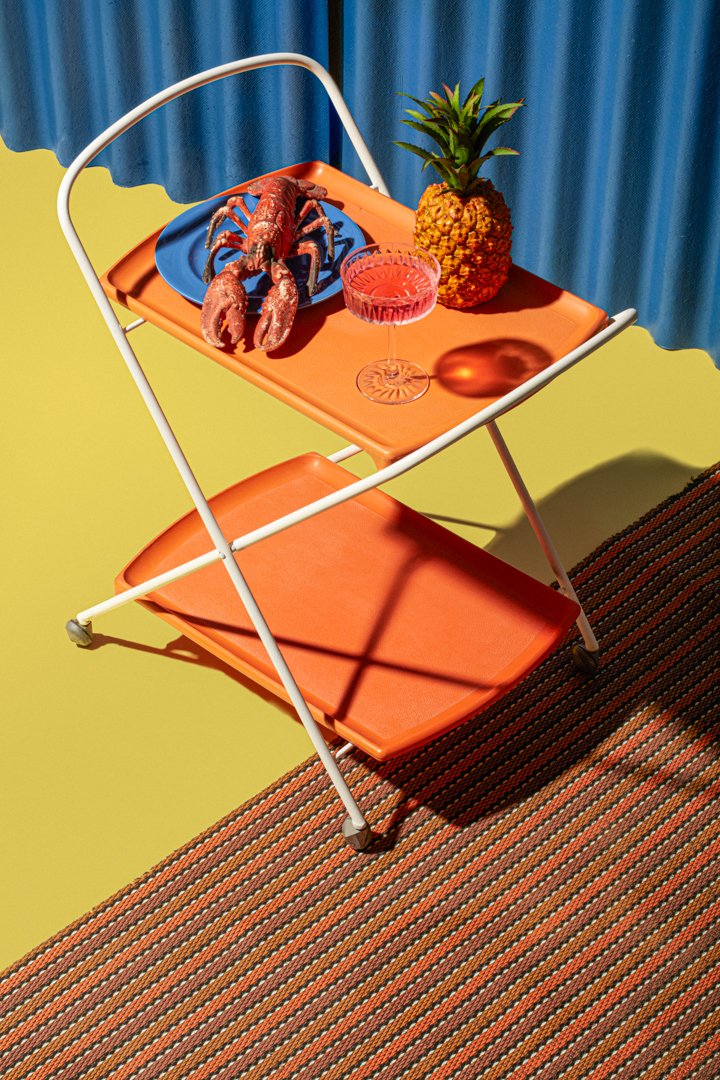 Two-tier orange serving trolley with a lobster on a plate, pineapple and pink cocktail, set on a striped rug