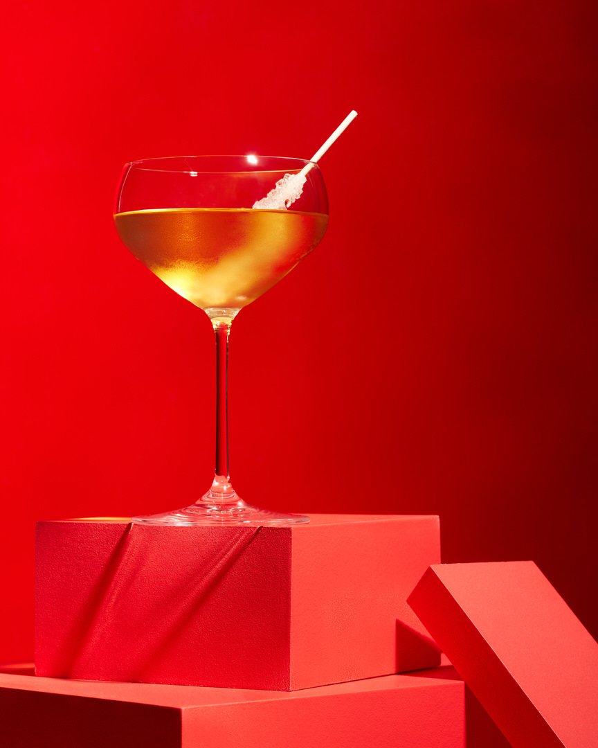 Golden cocktail in a coupe glass with a sugar-rimmed straw, set on red geometric blocks against a red backdrop