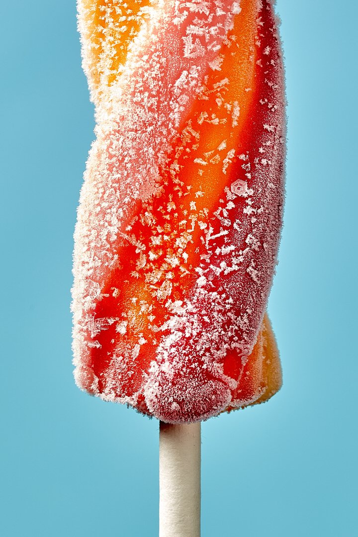 Close-up of a frosty orange and red ice lolly on a stick, covered in ice crystals, against a blue background