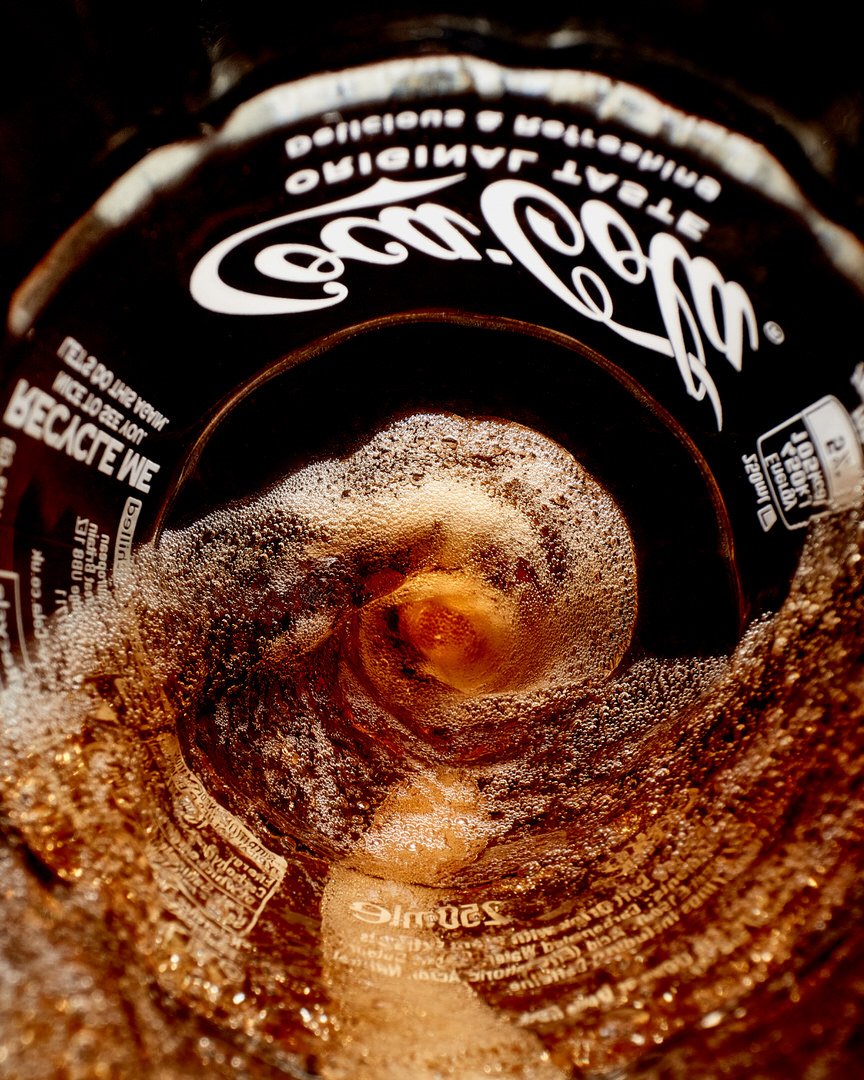 Close-up looking down into a fizzing Coca-Cola bottle, showing bubbles and the "Original Taste" label around the rim