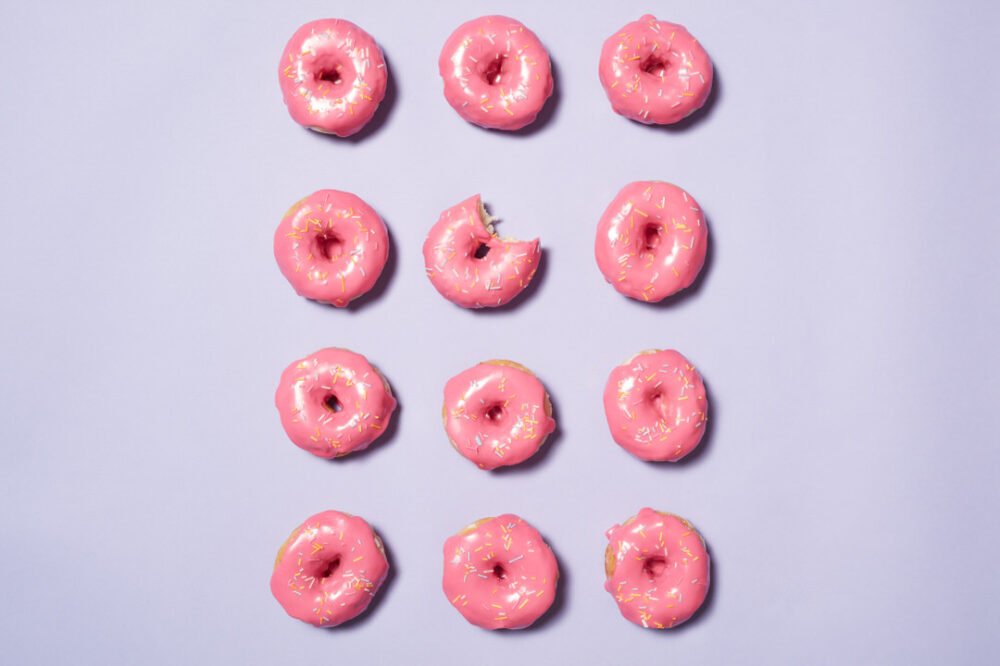 Grid of pink iced doughnuts with sprinkles on a lilac background, one with a bite taken out