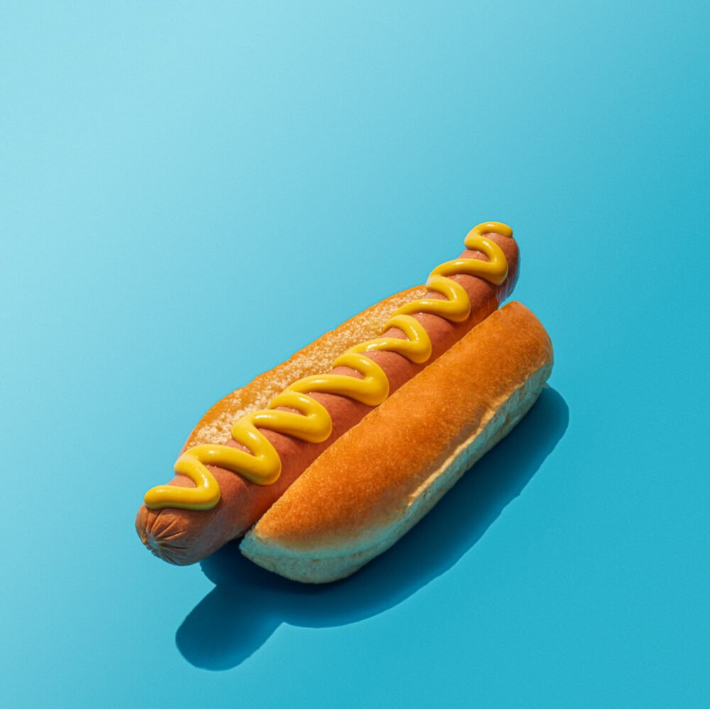 Hot dog in a bun topped with a zigzag of yellow mustard, on a blue background