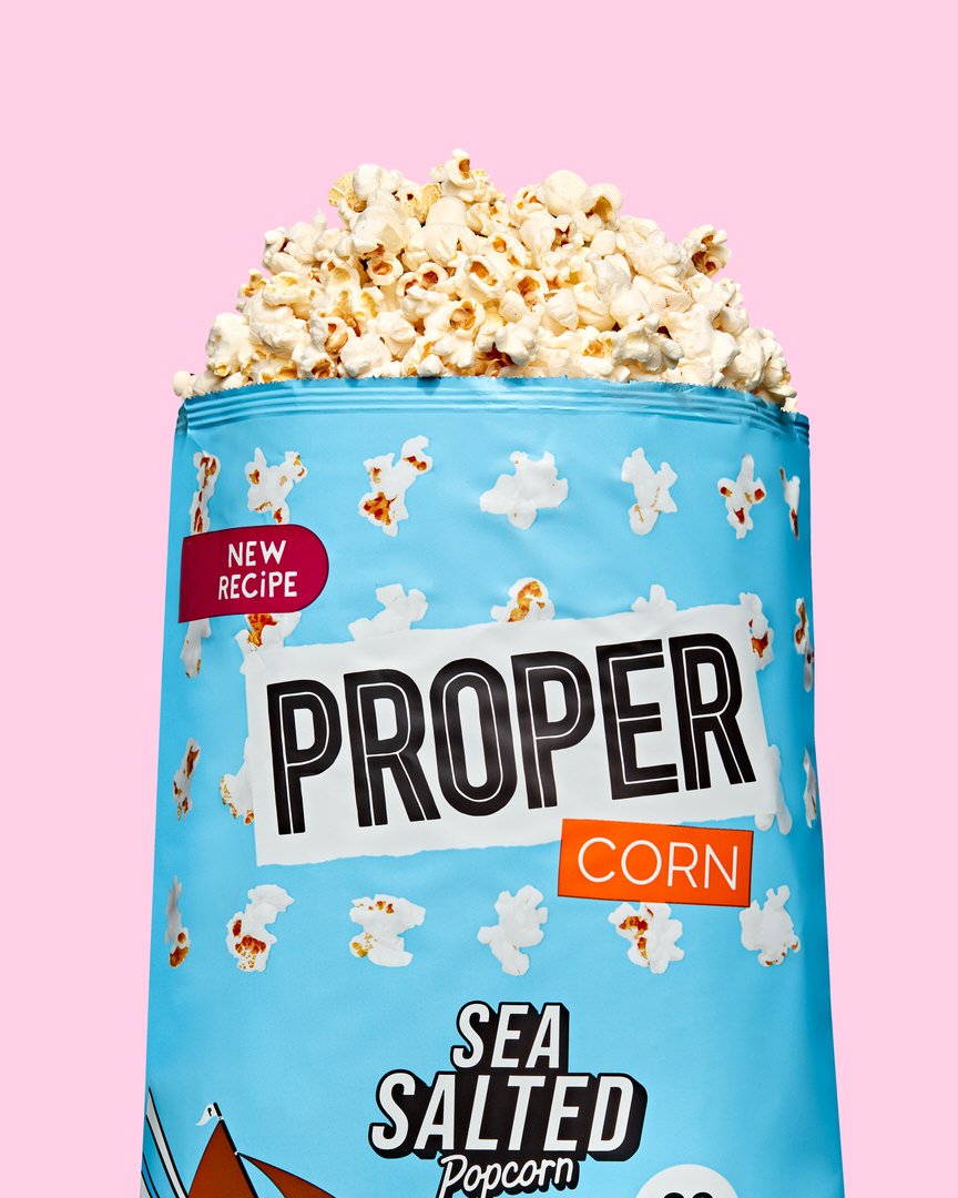 Blue Proper Corn Sea Salted Popcorn bag, marked "New Recipe", overflowing with popcorn