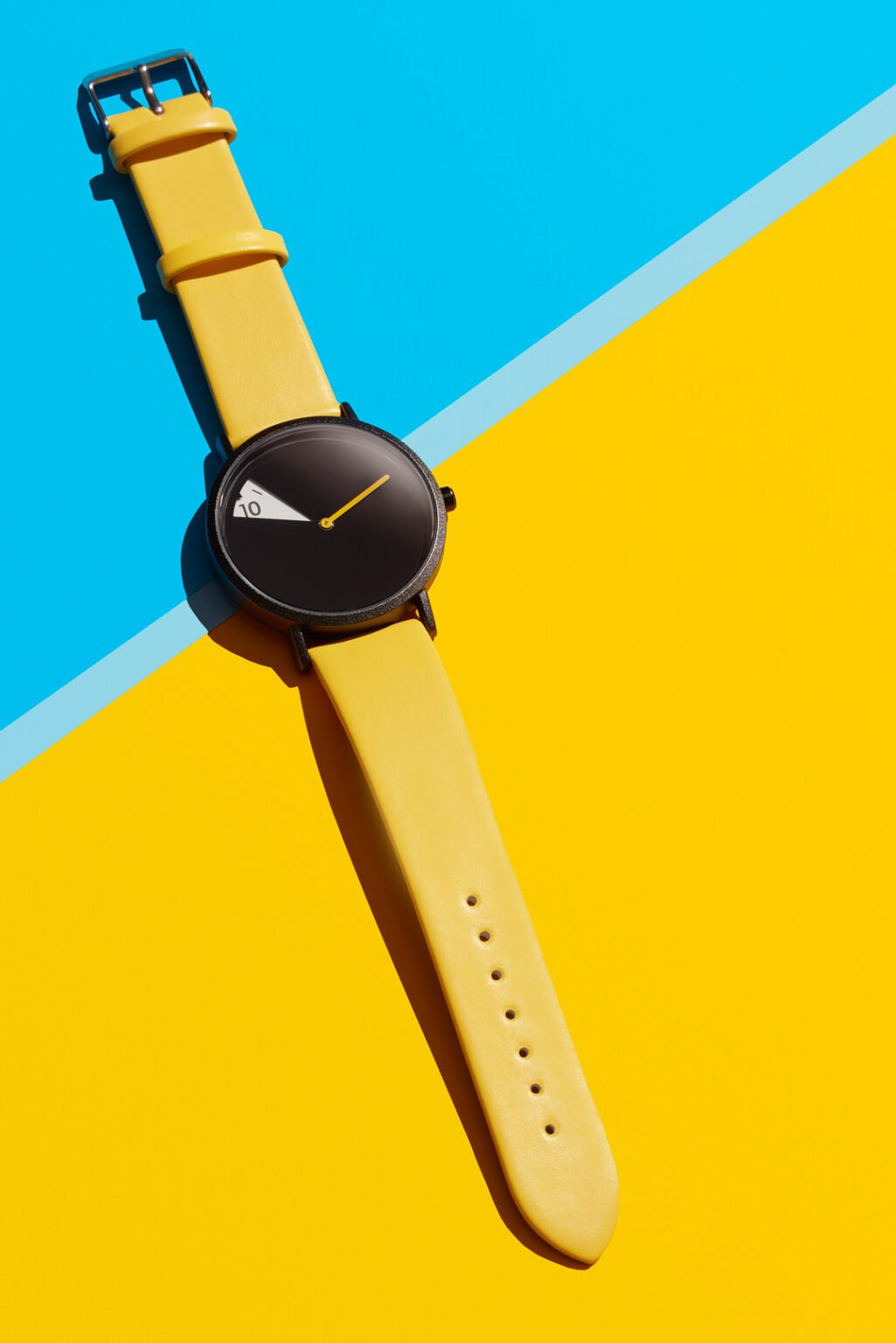 Watch with a black face and yellow leather strap, laid diagonally across a blue and yellow background