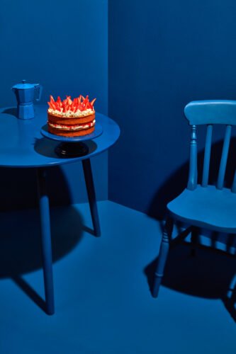 A strawberry layer cake and coffee pot on a table in a monochromatic blue room, with a chair beside it