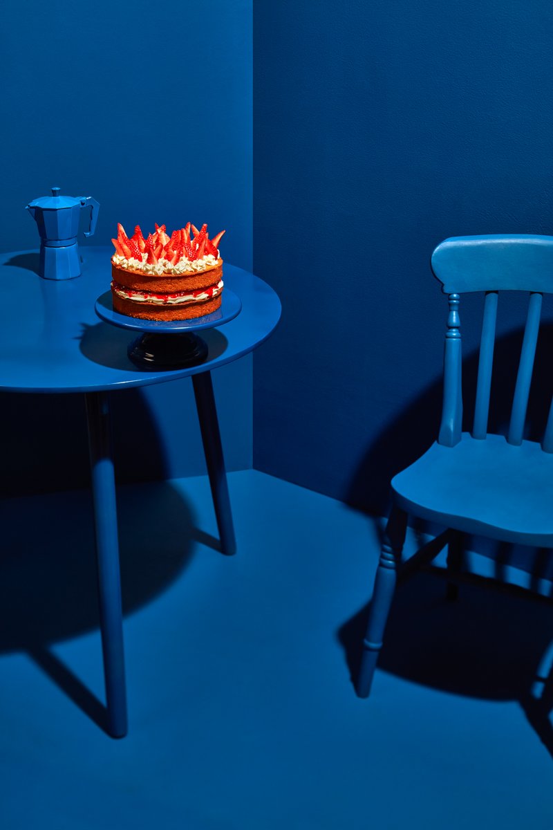 A strawberry layer cake and coffee pot on a table in a monochromatic blue room, with a chair beside it