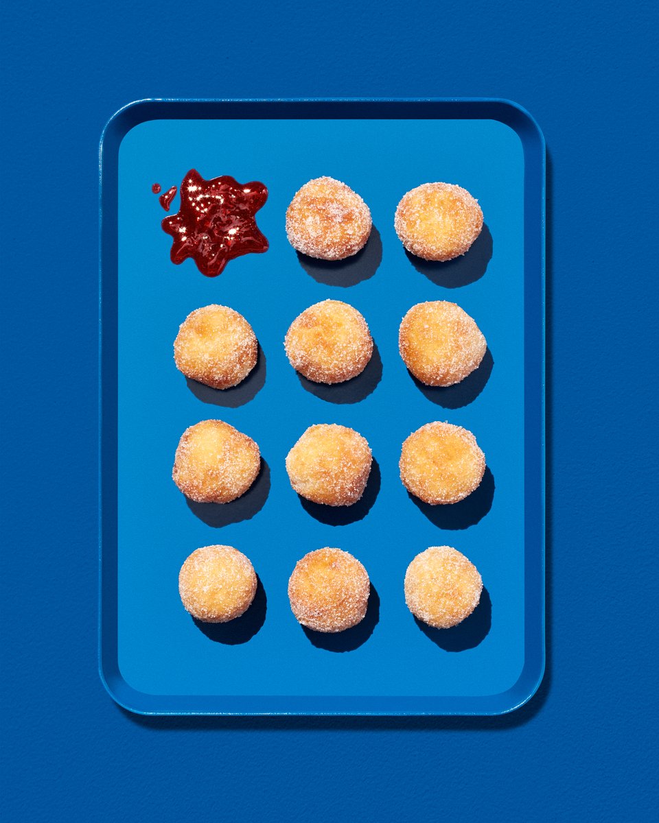 Rows of sugar-coated doughnut holes arranged on a blue tray, with a splash of red jam alongside them