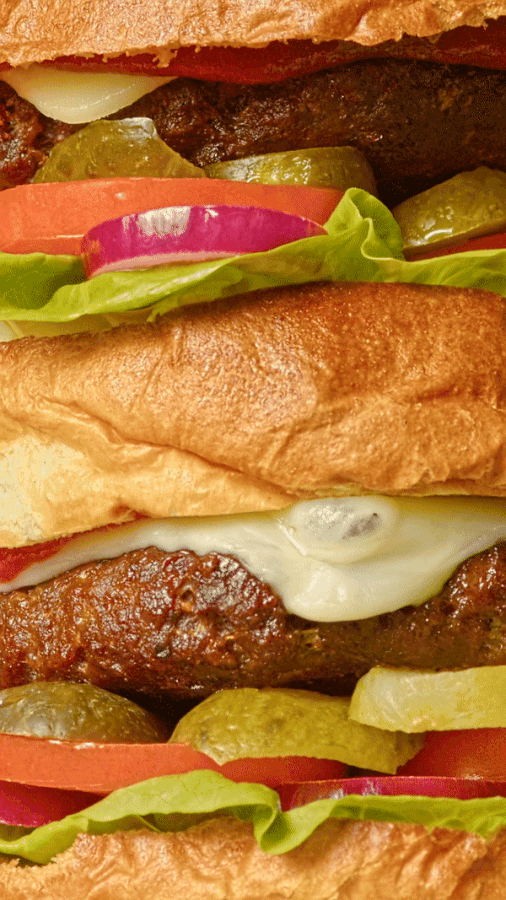 Close-up of stacked burgers with beef patties, melted cheese, pickles, tomato, red onion and lettuce between soft buns
