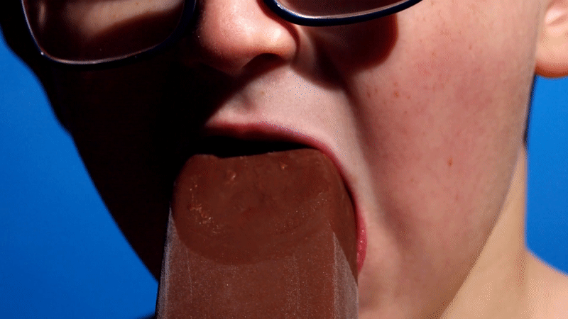 Close-up of a person wearing glasses licking a chocolate ice lolly
