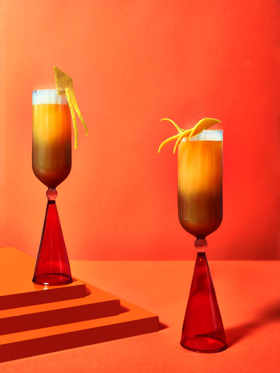 Two amber cocktails in tall red-stemmed glasses, garnished with citrus twists, on an orange background