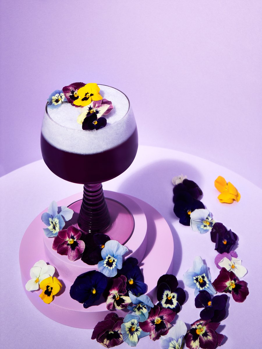 Purple frothy cocktail in a coupe glass topped with pansy flowers, surrounded by scattered pansies on a lilac table