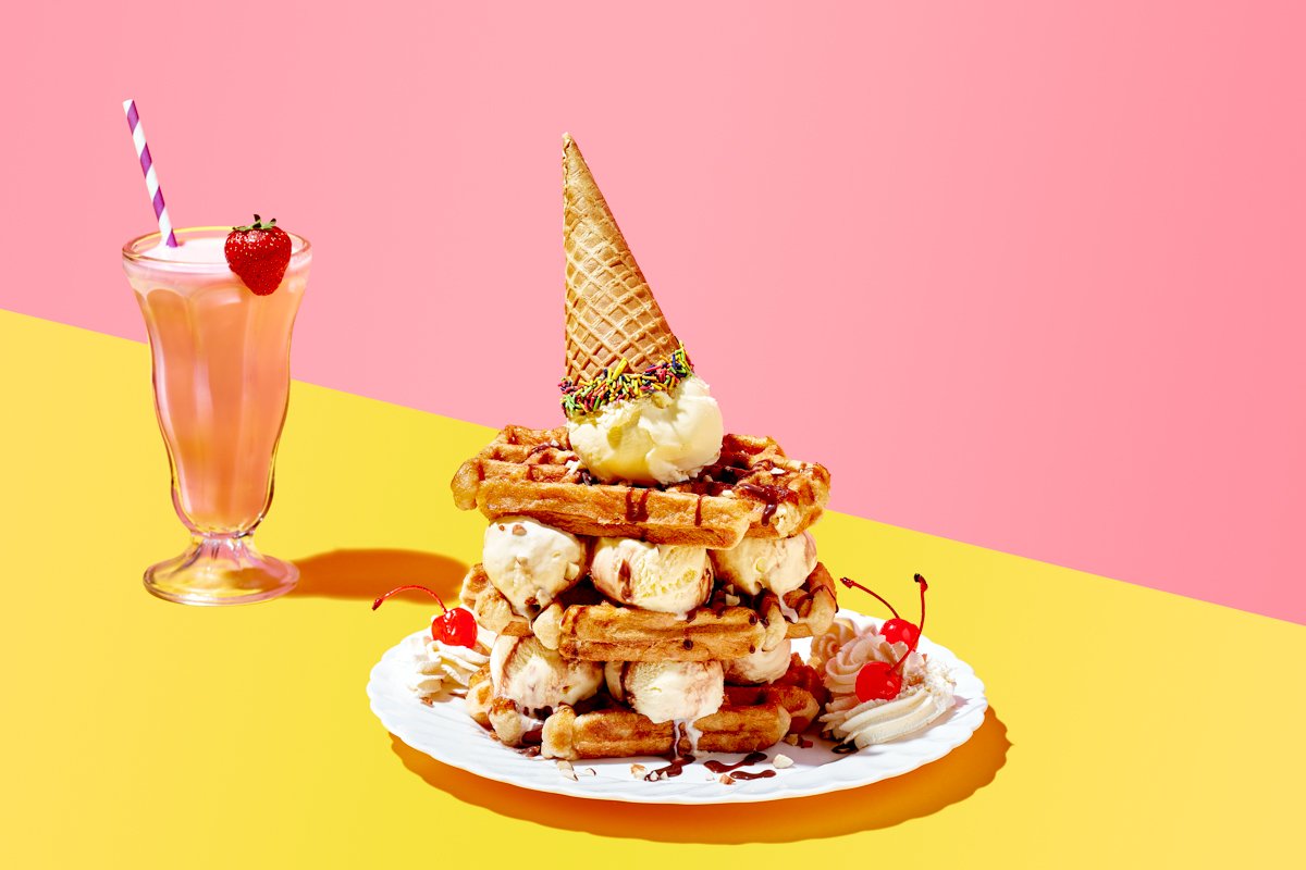 Stack of waffles layered with ice cream scoops, topped with an upturned ice cream cone, served with cherries, cream and a strawberry milkshake