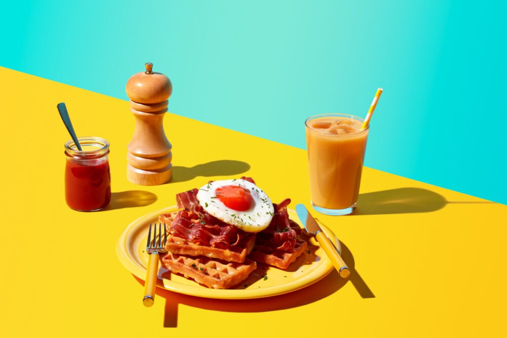 Waffles topped with a fried egg and bacon, served with a jar of ketchup, a pepper grinder and an iced drink