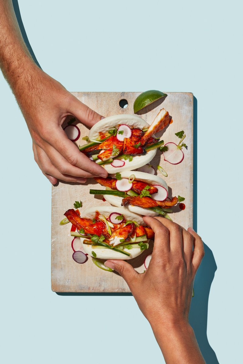 Hands picking up bao buns filled with glazed chicken, cucumber, radish and coriander on a wooden board
