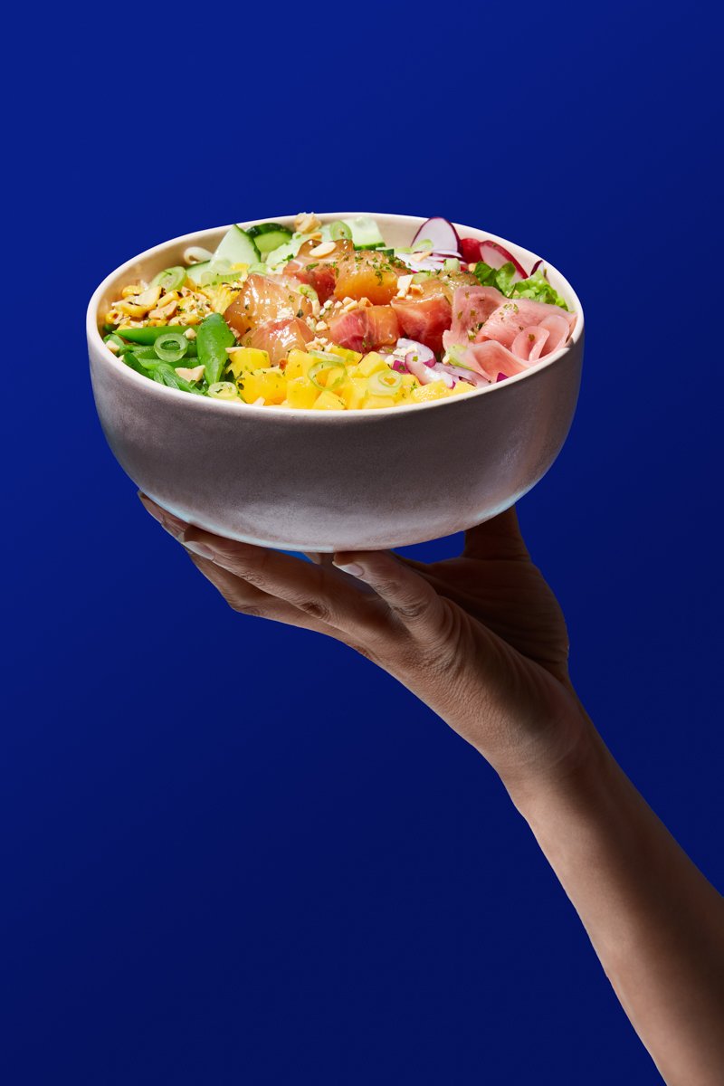 A hand holding a bowl of poke salad with salmon, mango, radish, pickled ginger and herbs against a blue background