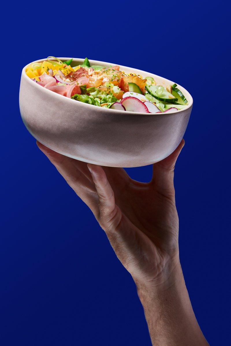 A hand holding up a bowl of poke-style salad with mango, cucumber, radish, salmon and pickled onion, against a blue background