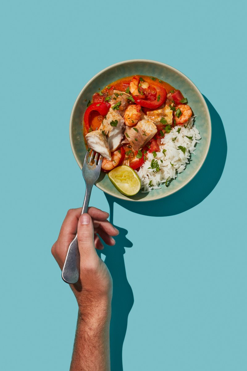 A hand holds a fork up to a bowl of fish and prawn stew with rice, peppers and lime, on a teal background