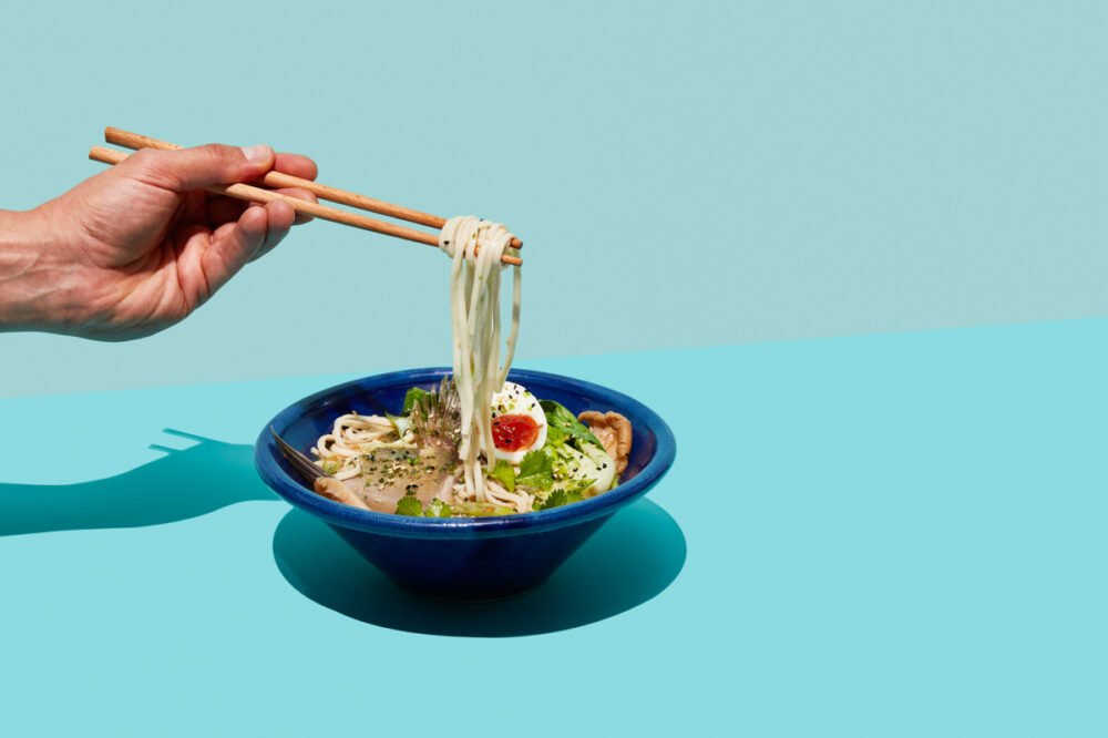 A hand lifts noodles with chopsticks from a blue bowl of ramen with egg, greens and mushrooms