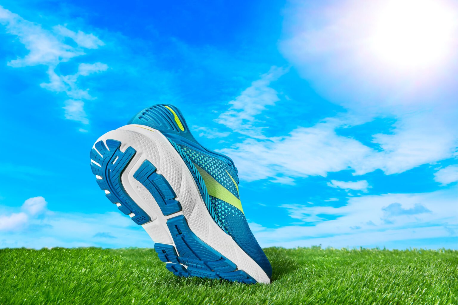 Blue and white running shoe with yellow accents, standing upright on grass against a bright blue sky, showing its tread pattern