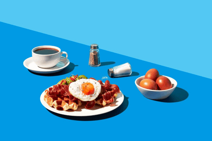 Breakfast spread on blue background: coffee cup, salt and pepper shakers, a plate of waffles with bacon, a fried egg and salad garnish, and a bowl of eggs