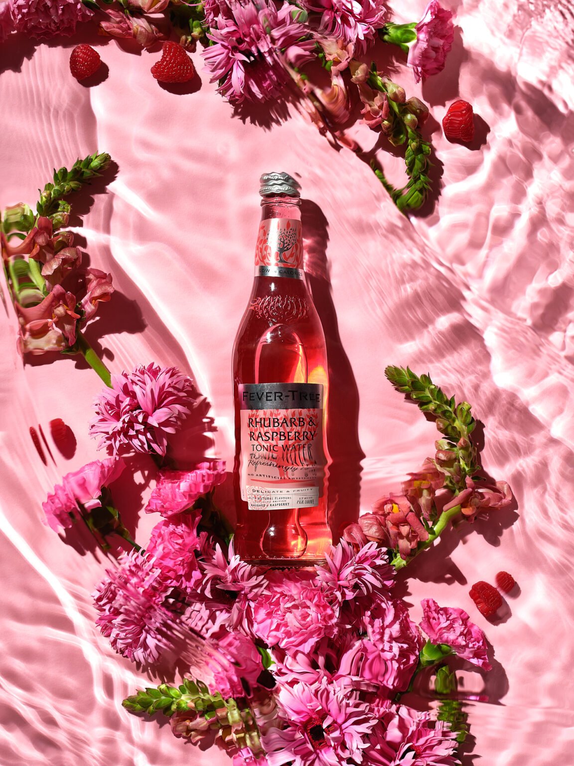 Bottle of Fever-Tree Rhubarb & Raspberry Tonic Water surrounded by pink flowers and raspberries on a pink water backdrop