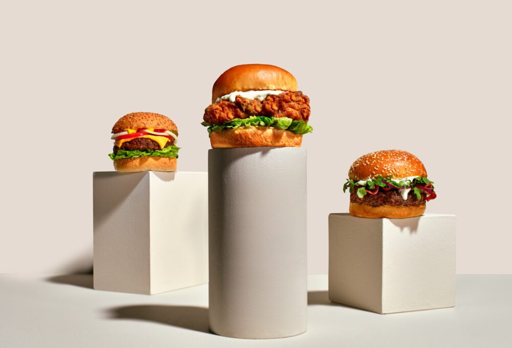 Three burgers displayed on plinths of varying heights: a classic cheeseburger, a crispy chicken burger, and a burger topped with mixed leaves and slaw
