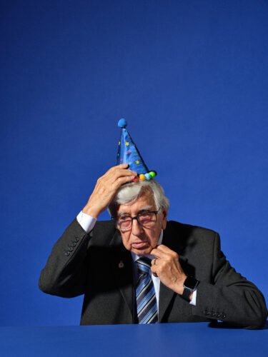 advertising photograph of an old man adjusting his party hat taken on a blue studio background in london