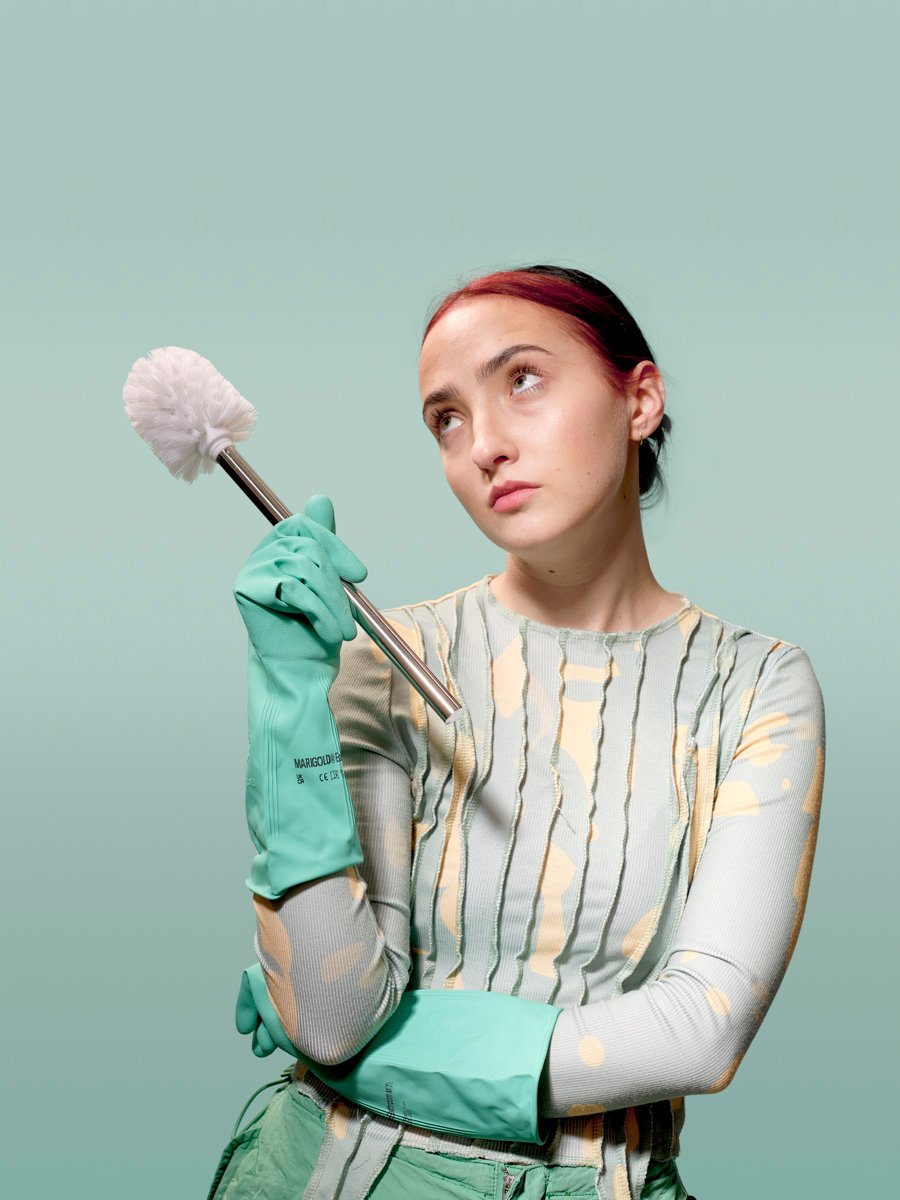 Woman wearing rubber gloves holds a toilet brush aloft, gazing upward with a wistful expression