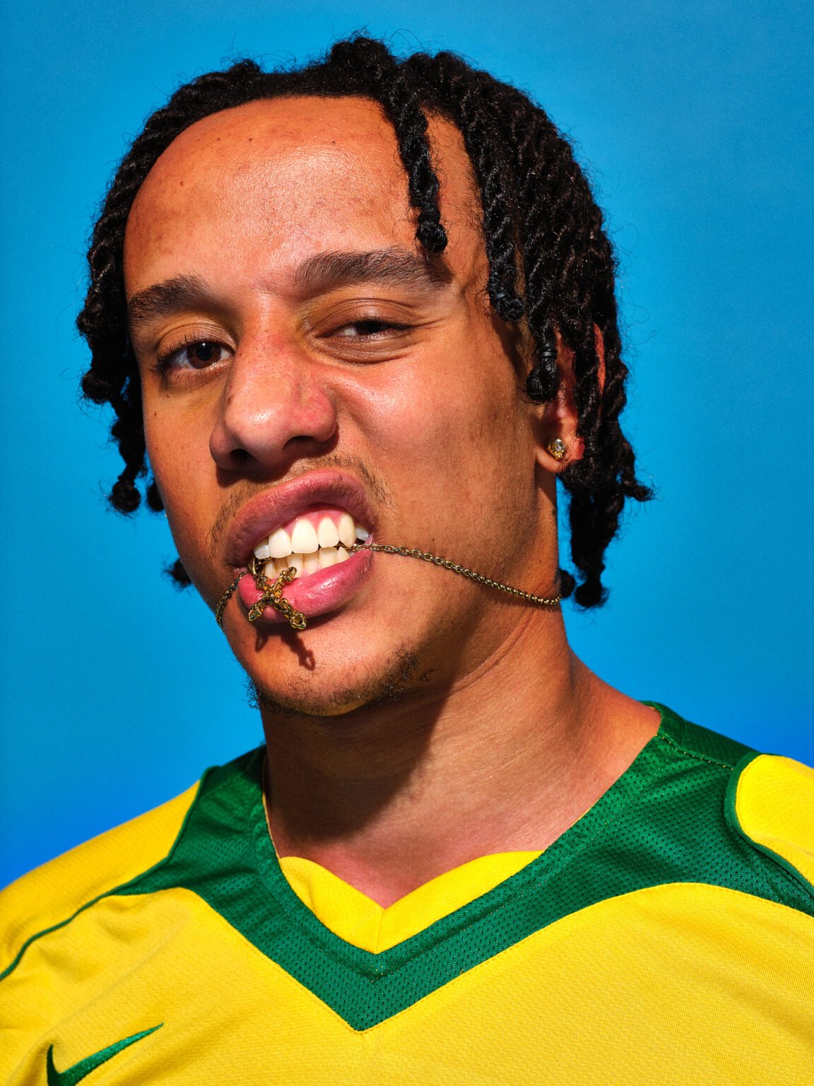 Man with braided hair wearing a yellow and green shirt, biting a gold cross pendant on a chain