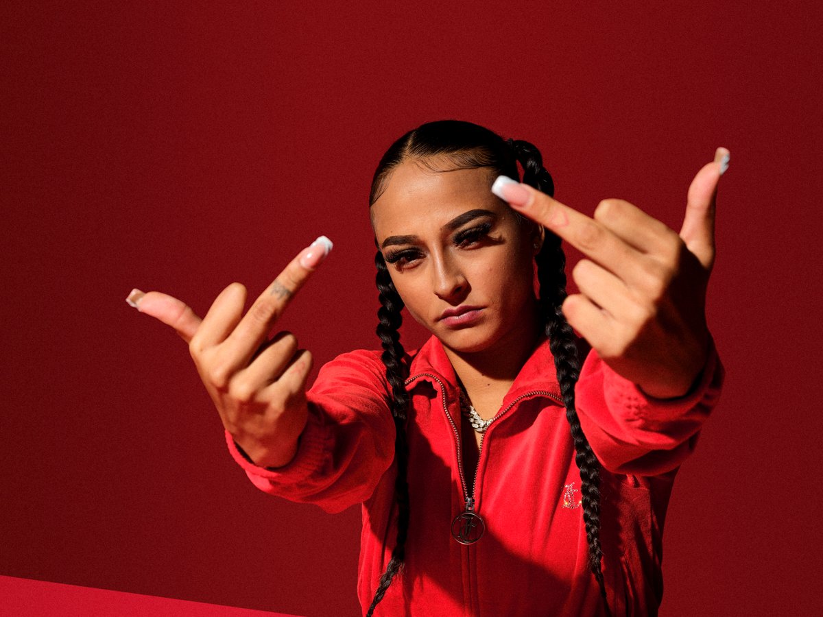 Woman with braided hair in a red tracksuit, raising both middle fingers towards the camera against a red backdrop