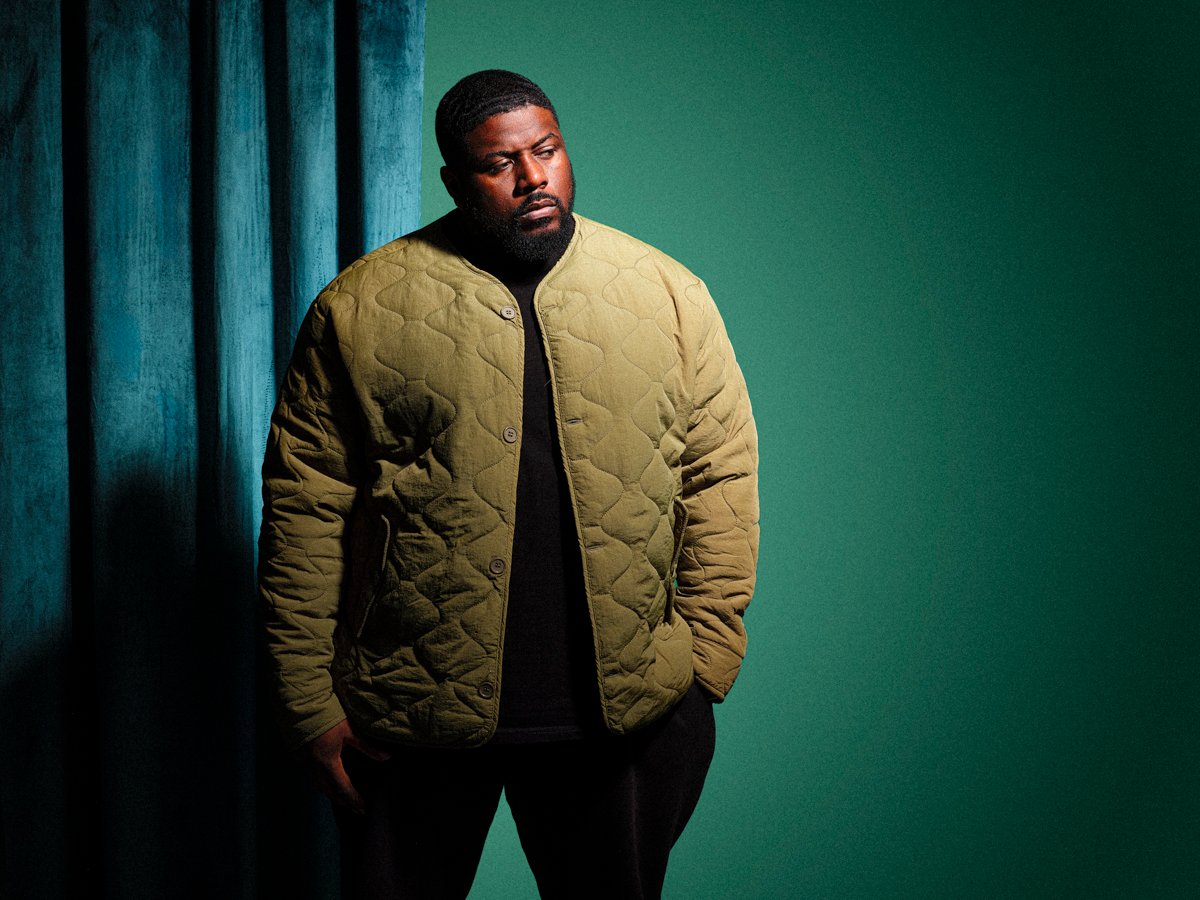 Man with a beard wearing an olive quilted jacket over a black T-shirt, standing beside a blue curtain
