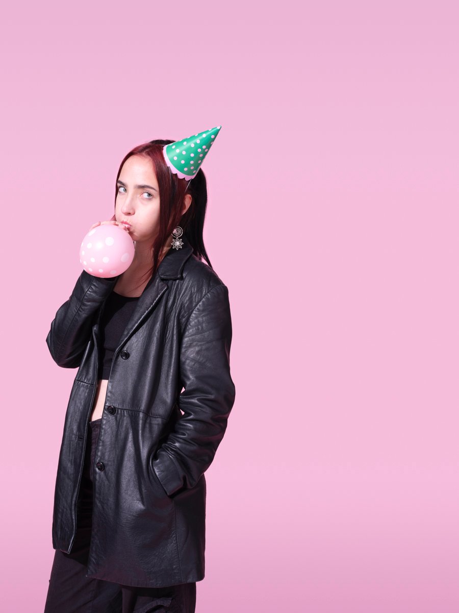 Woman in a black leather coat and party hat blowing up a polka-dot balloon