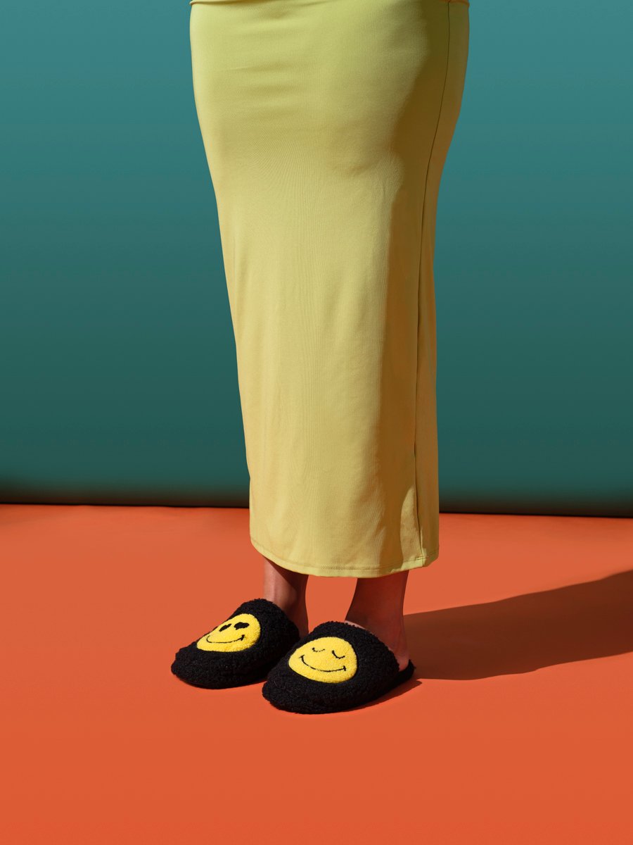Person wearing a yellow midi skirt with black fluffy slippers featuring embroidered smiley faces