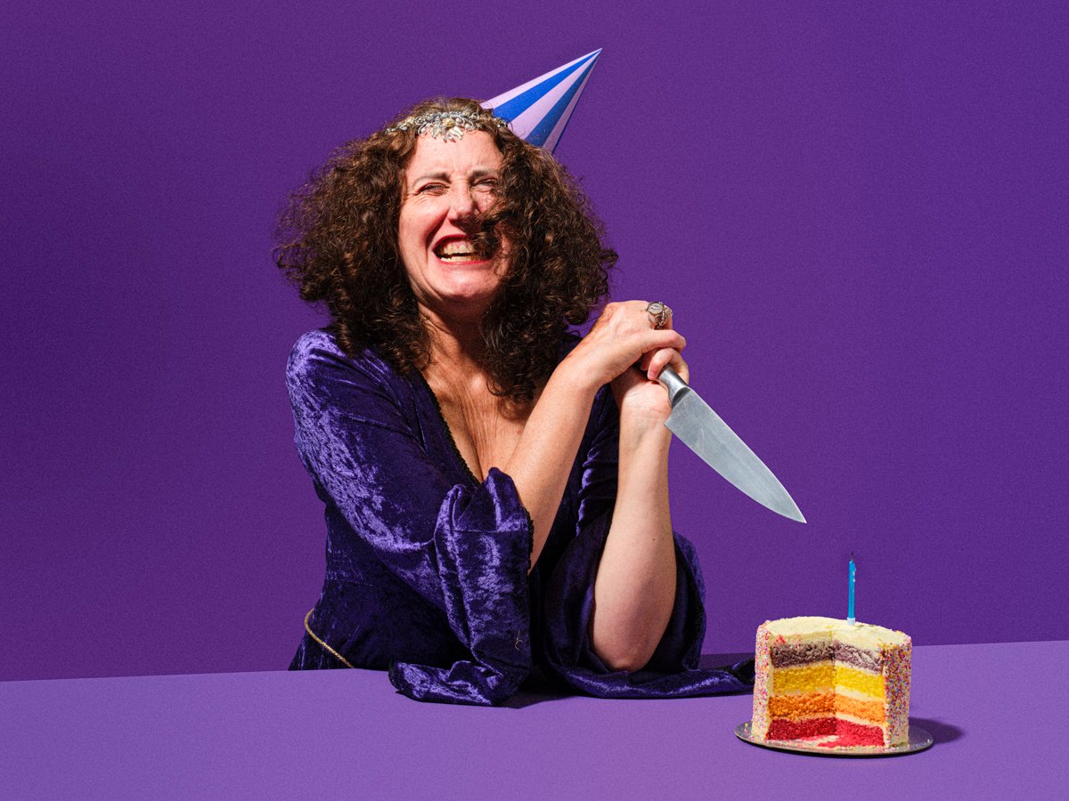 a middle aged lady in purple dress on a purple photo studio background stabbing a cake