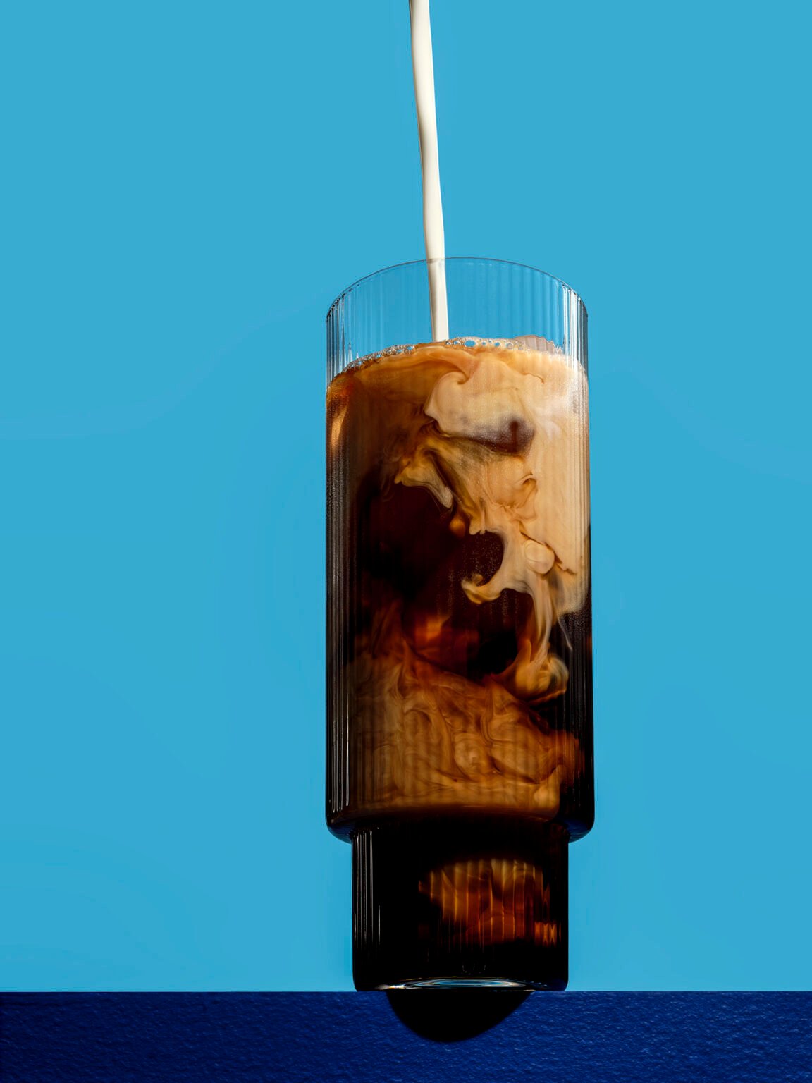hero drink photograph of an iced coffee on a blue background with the milk perfectly swirling around the glass