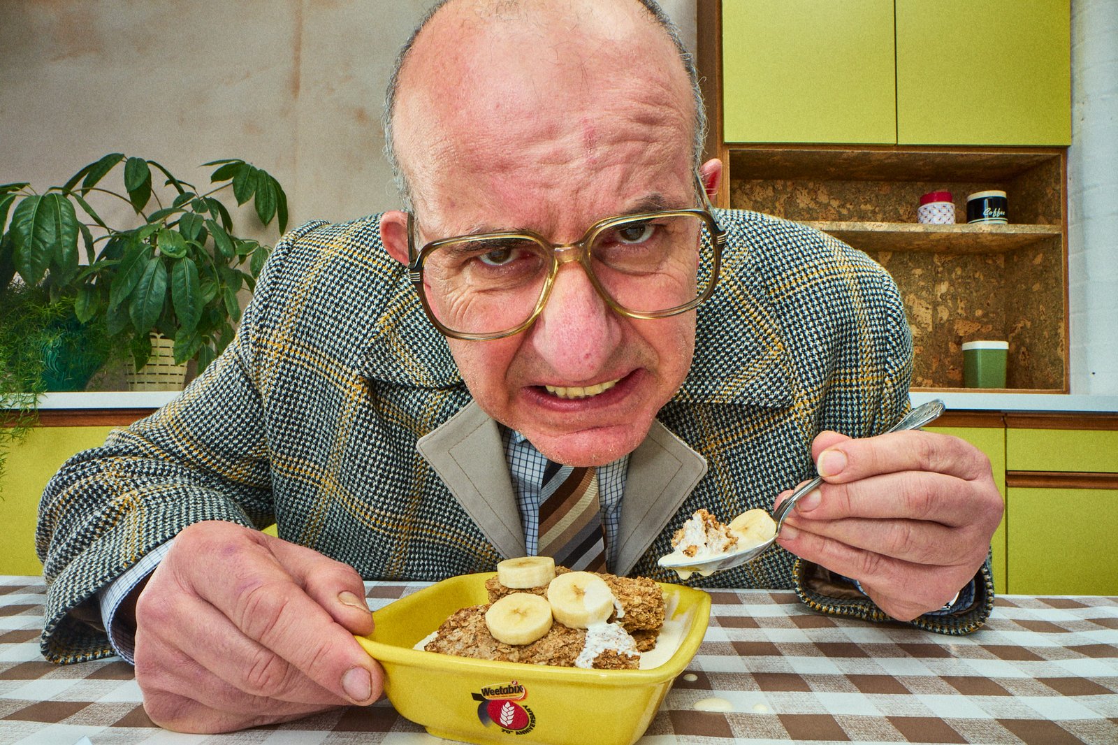 Bald man in tweed jacket pulling an angry face while eating Weetabix with banana slices from a yellow bowl