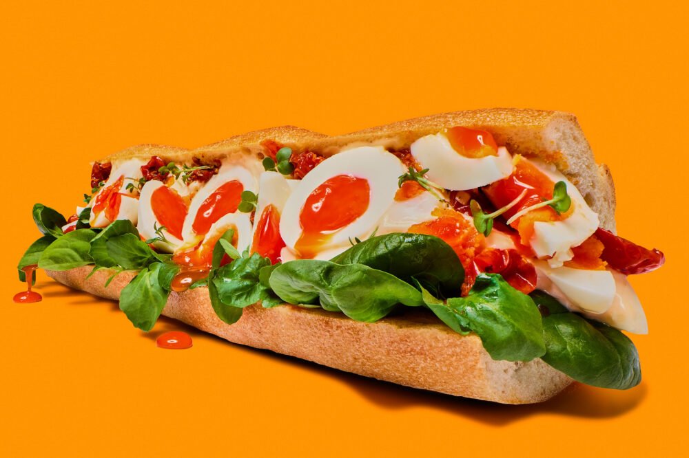 a bold and graphic advertising photograph of an over the top egg baguette on a bright orange background