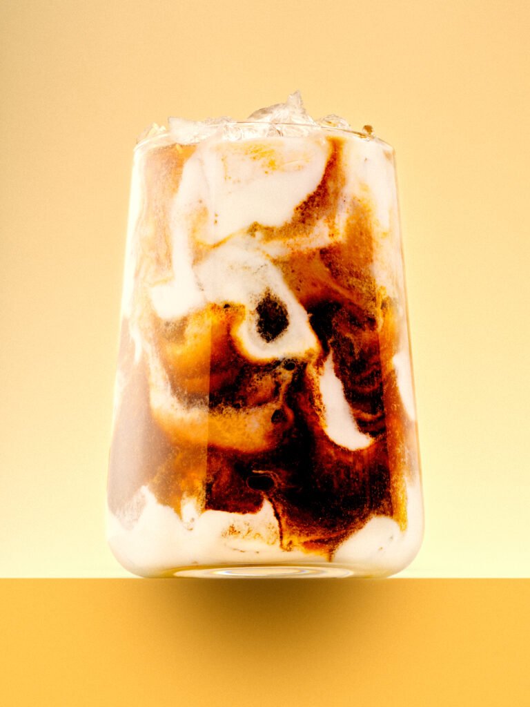 Close-up of an iced coffee drink with swirling milk and dark coffee layers, ice on top, against a warm yellow background