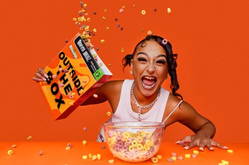 Woman laughing while pouring "Outside The Box" cereal into a bowl, orange backdrop